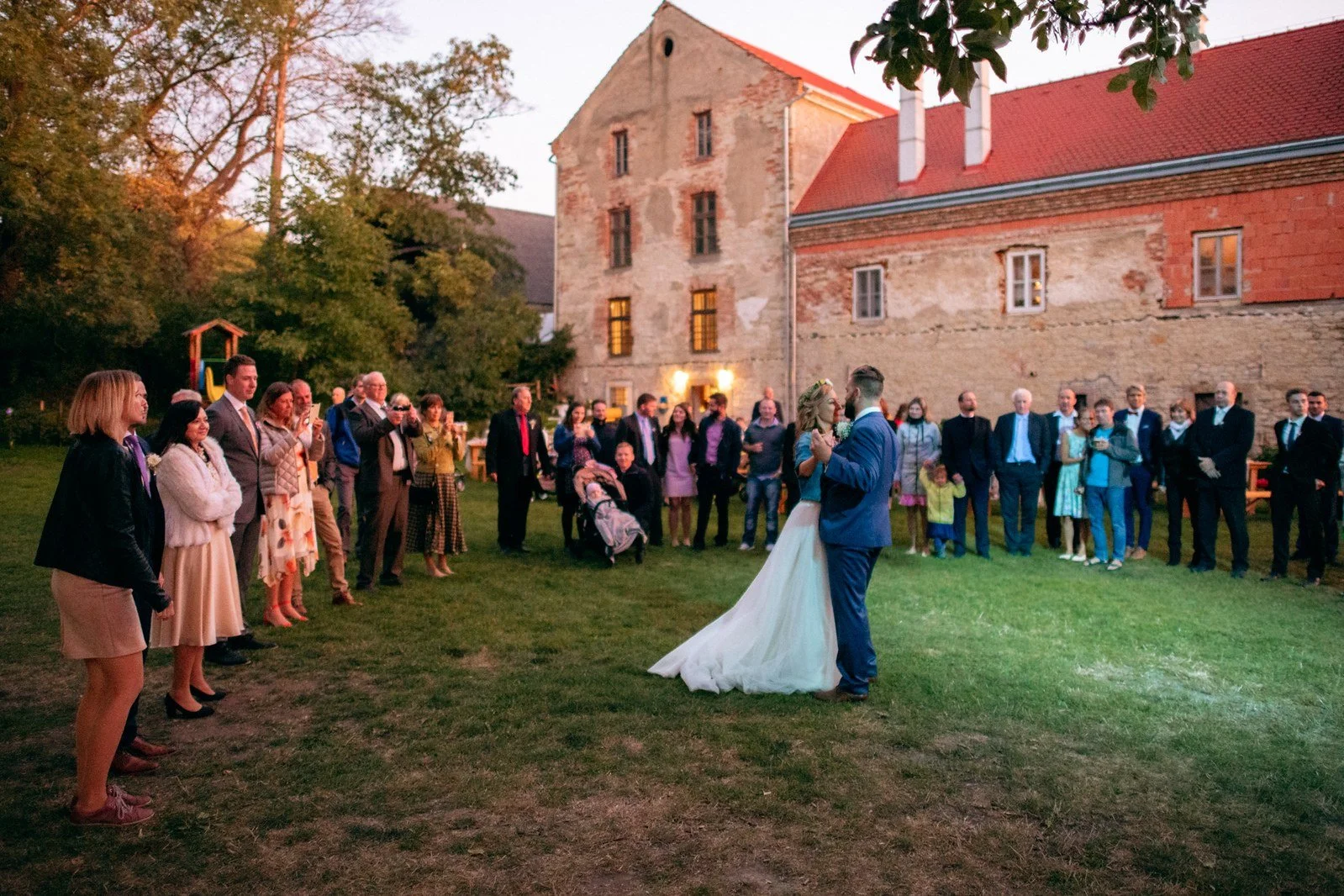 Skupina hostů se shromažďuje kolem ženy v bílých šatech na dvoře před cihlovou budovou.
