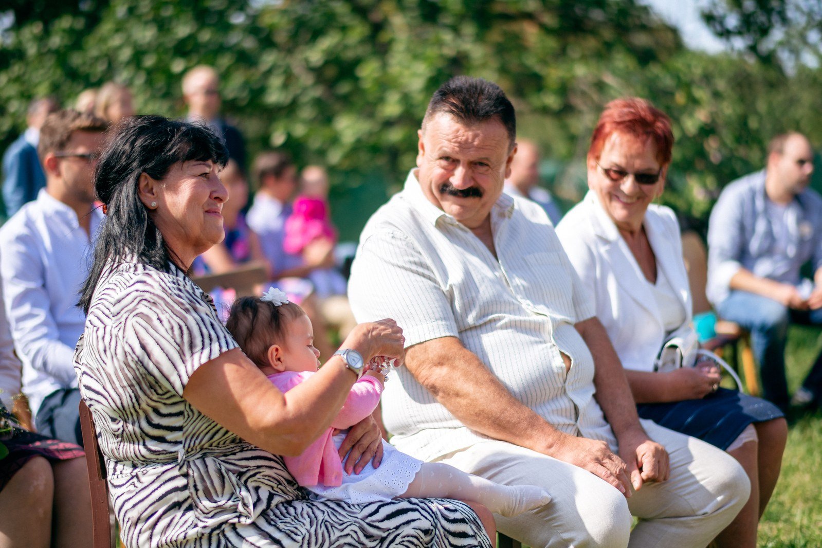 Sedící žena v pruhovaných šatech drží za ruku muže s knírem, vedle něhož sedí žena s krátkými zrzavými vlasy.