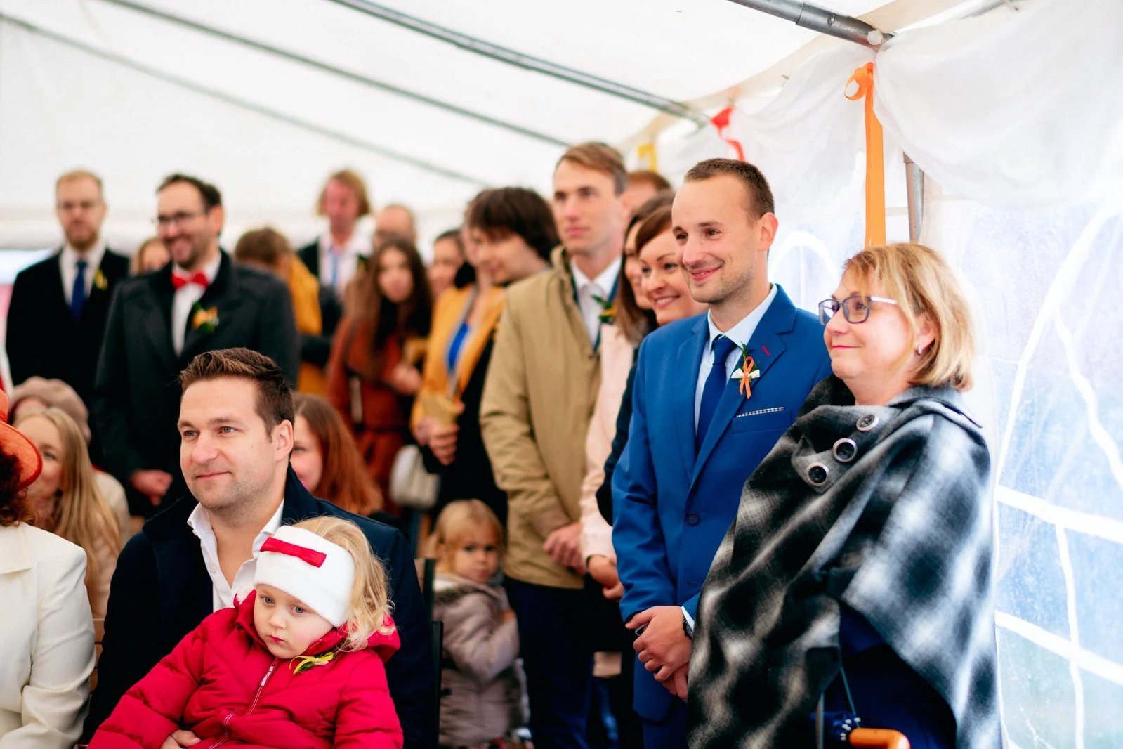 Muž v modrém obleku stojí mezi hosty, z nichž část sedí a část přihlíží.