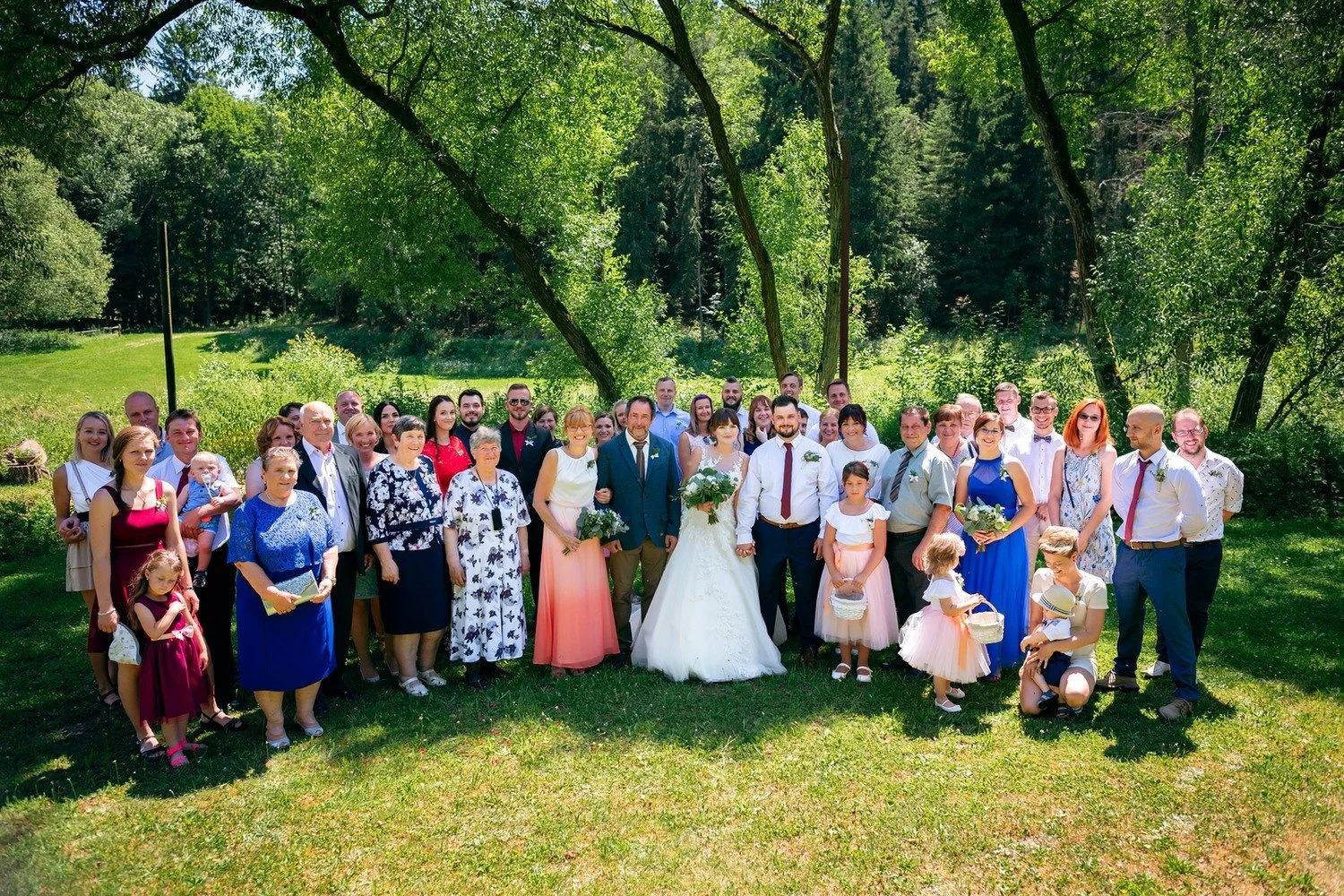 Velká skupina svatebních hostů pózuje společně na trávníku pod rozložitými stromy.