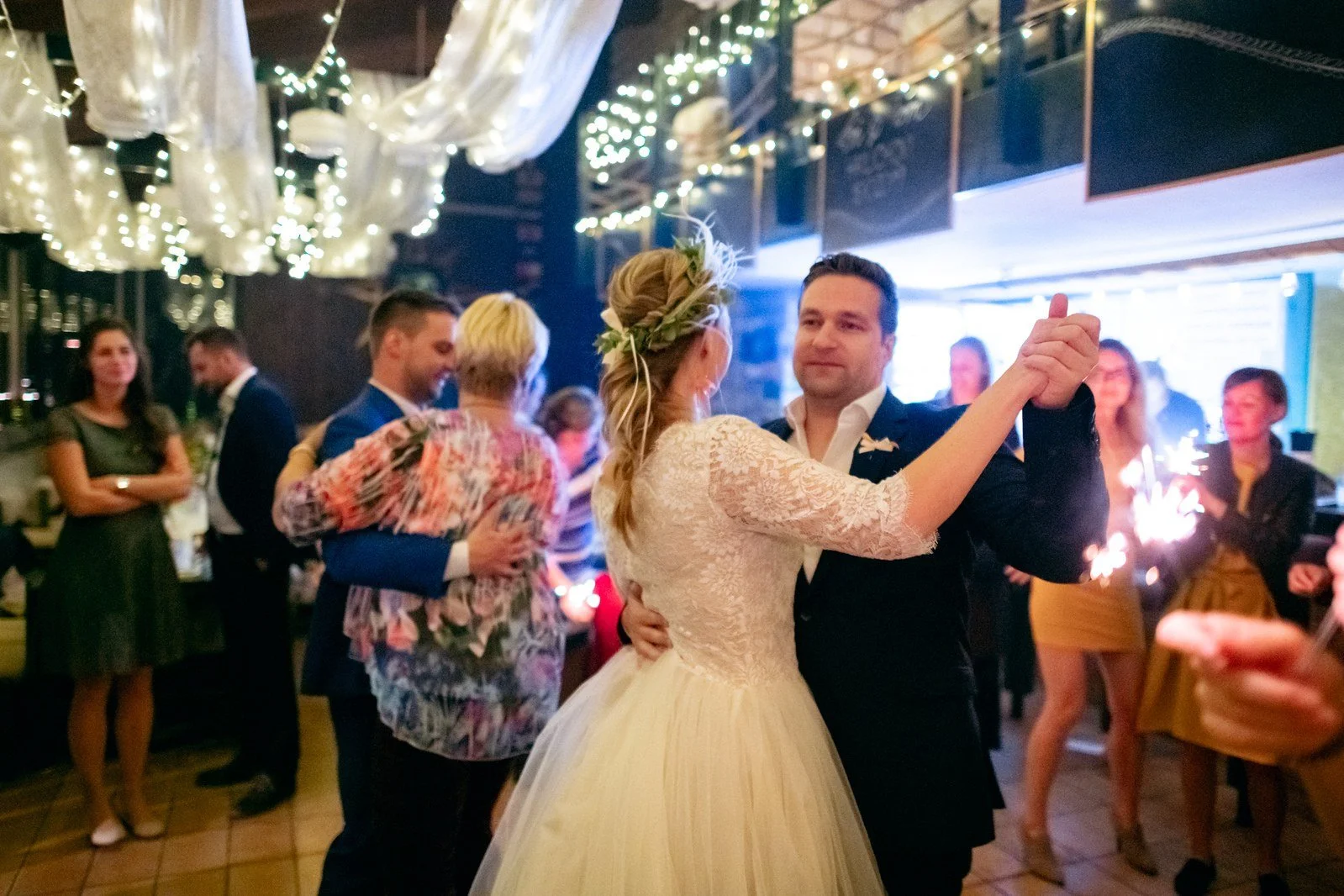 Žena v bílých šatech tančí s mužem ve světlé košili před přihlížejícími hosty.