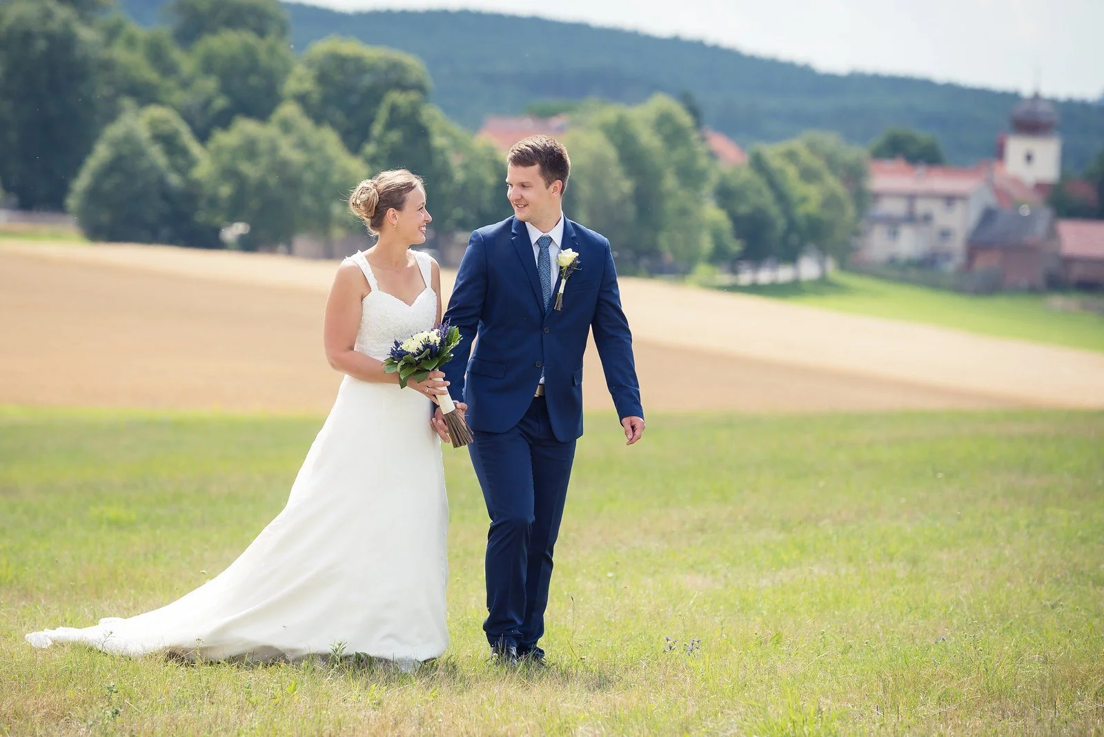 Nevěsta a ženich jdou ruku v ruce po okraji pole.