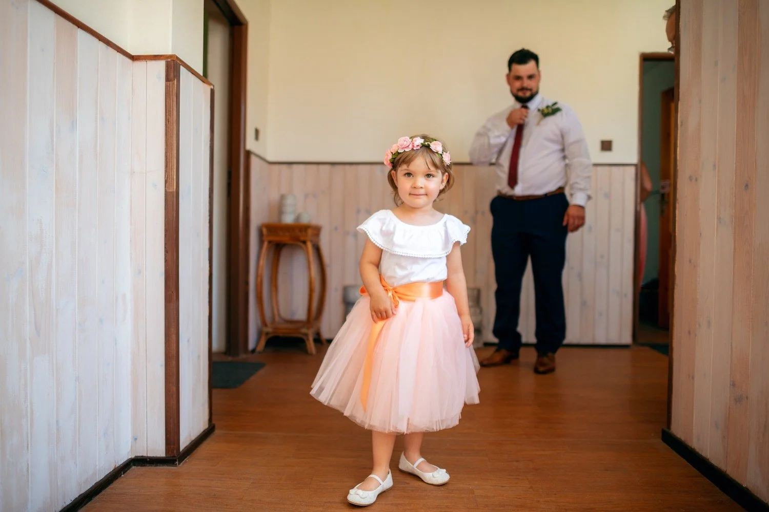 Dívka v růžové tylové sukni kráčí chodbou k fotoaparátu, zatímco Pavel stojí ve dveřích za ní.