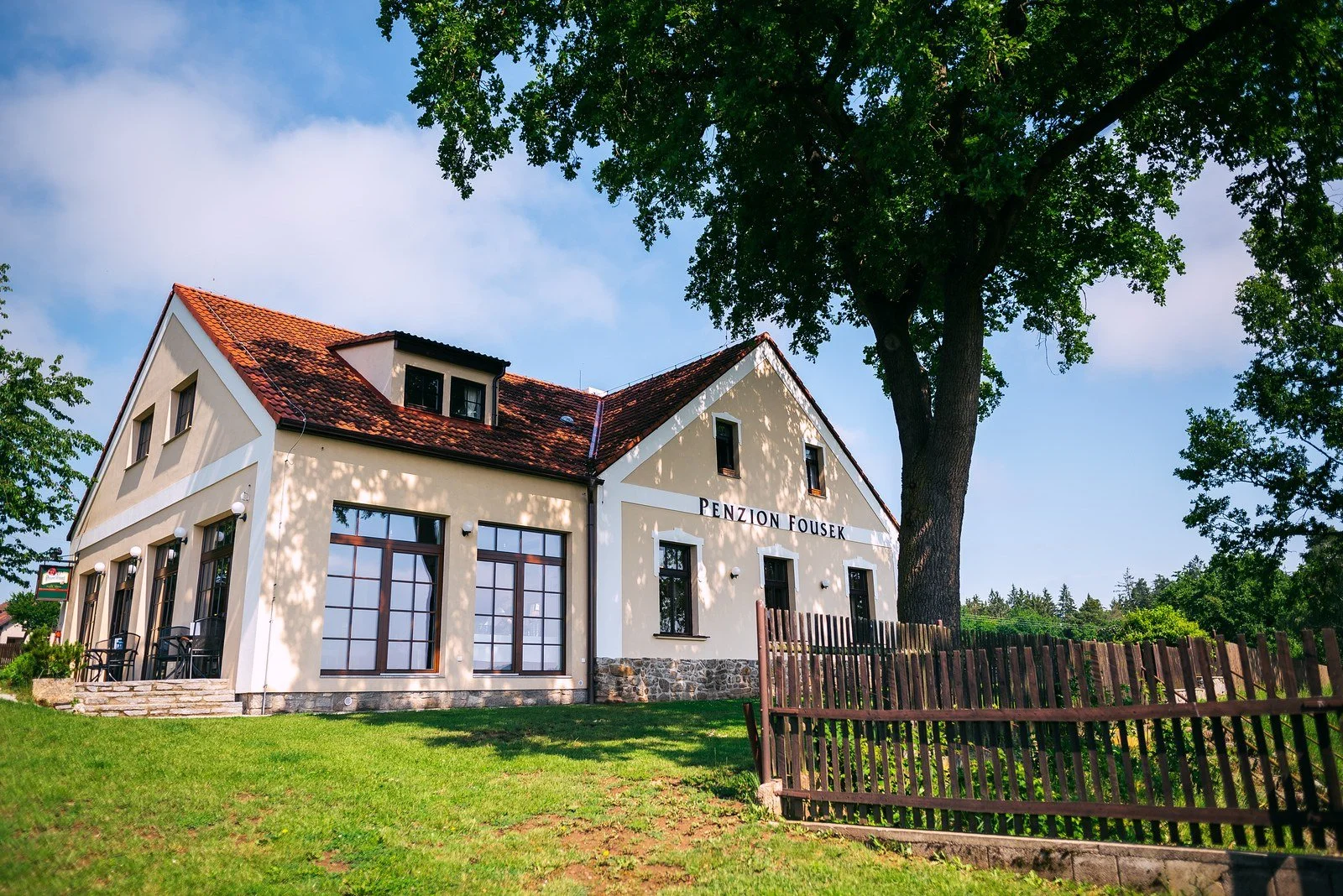 Historická budova s červenou střechou stojí za dřevěným plotem pod korunou stromu.