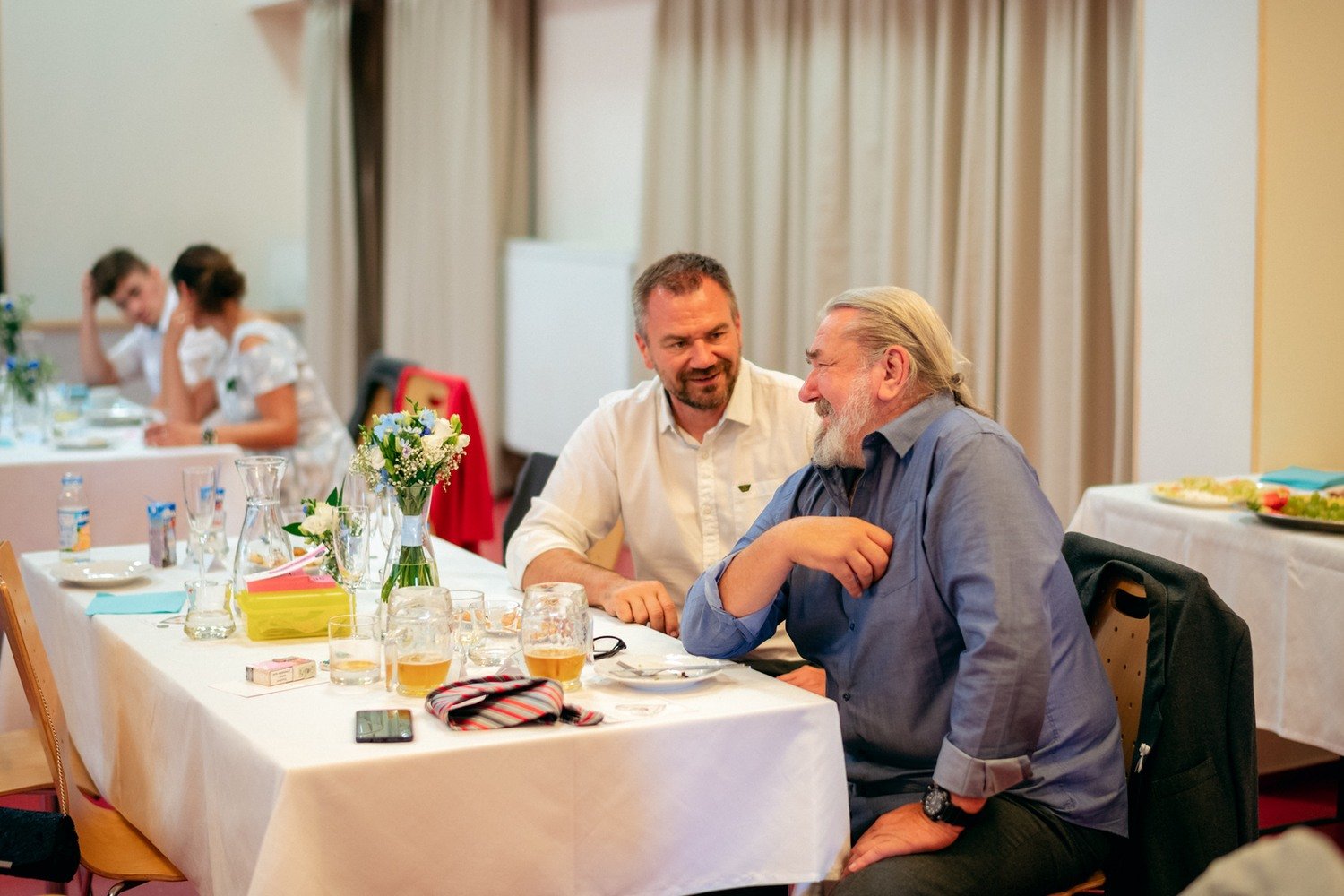 Dva muži spolu hovoří u prostřeného stolu v jídelním sále.