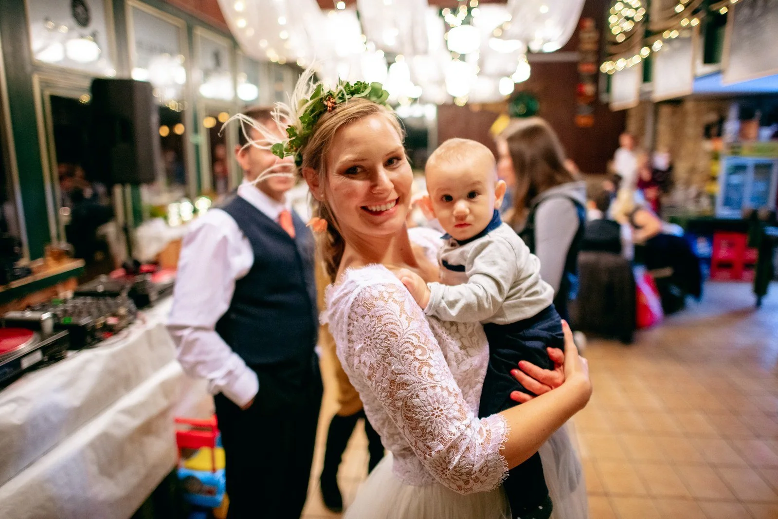 Žena v bílých krajkových šatech se sklání k jiné ženě v objetí mezi hosty.