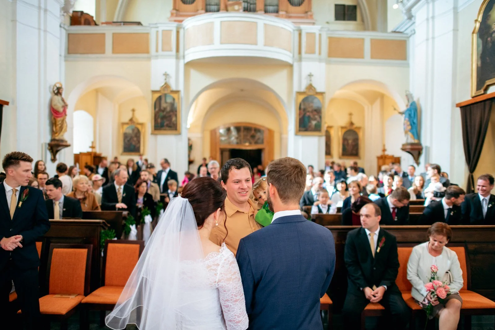 Dvojice ve svatebním oděvu stojí zády k fotoaparátu před mužem v košili a zaplněnými lavicemi.