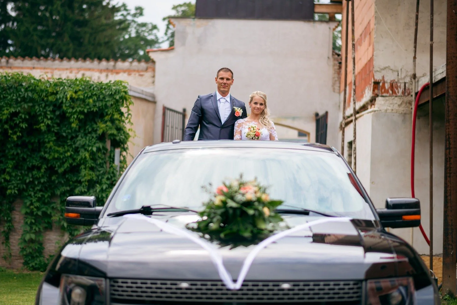 Jitka s Lukášem se usmívají nad střechou černého vozu s květinovou výzdobou na kapotě.