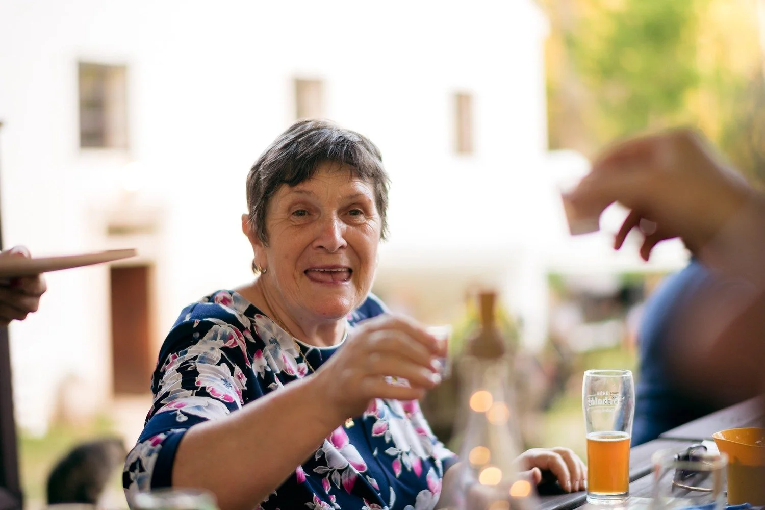 Žena v květované halence zvedá skleničku a pobaveně reaguje u venkovního stolu.