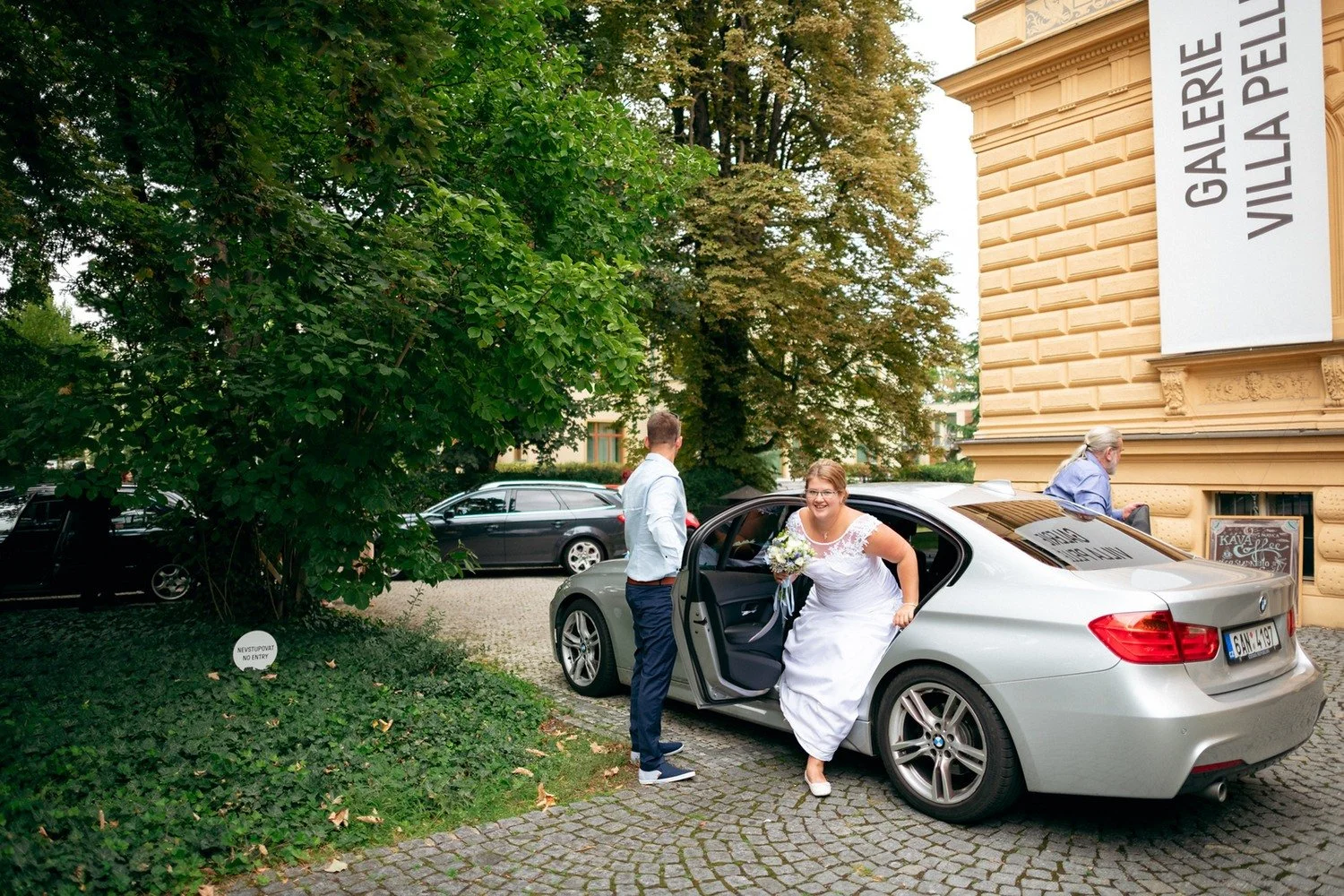 Žena v bílých šatech vystupuje z auta před Galerií Villa Pellé.