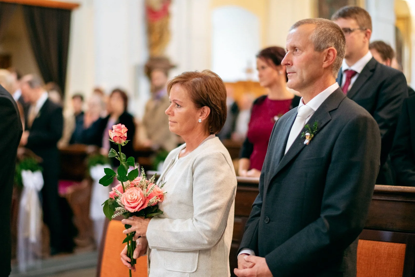 Žena ve světlém kostýmku drží růžové květy vedle muže v tmavém obleku v kostelní lavici.