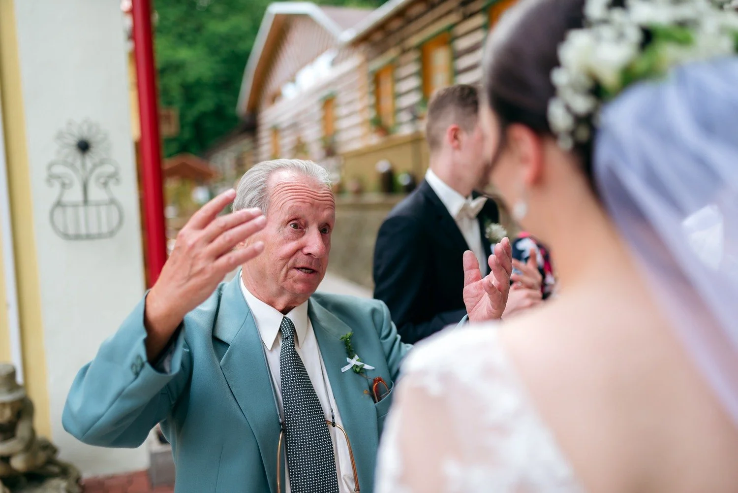Starší muž ve světle modrém saku živě gestikuluje při rozhovoru s nevěstou a ženichem.