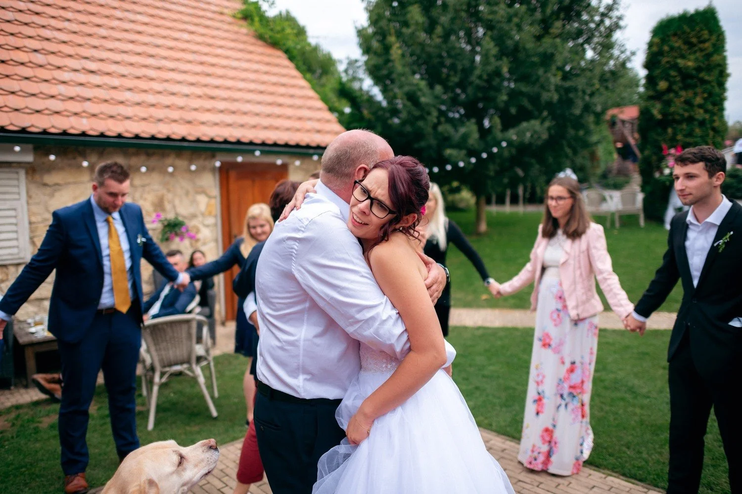 Muž v bílé košili pevně objímá ženu v bílých šatech během tance.