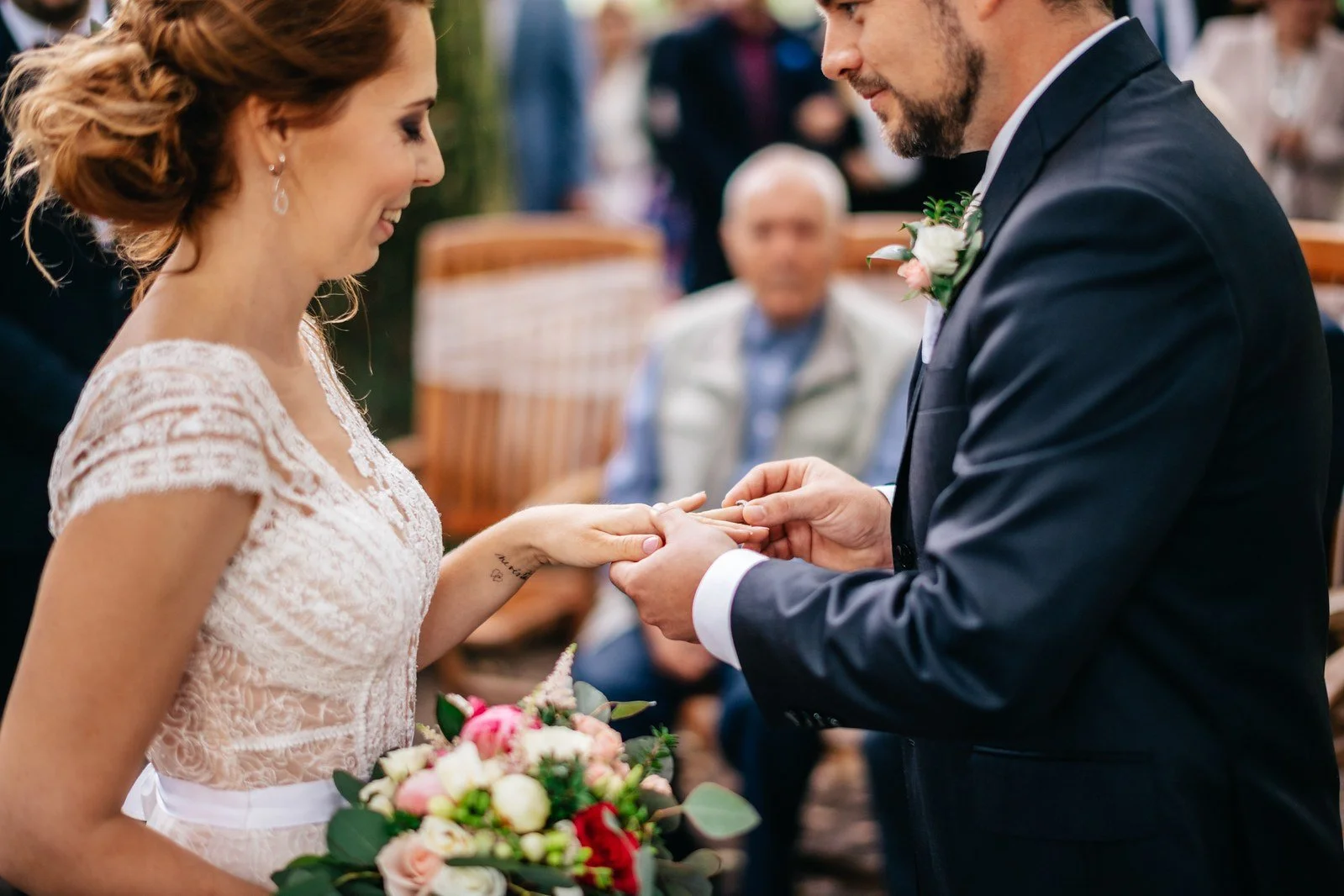 Detail dvojice ukazuje muže v tmavém obleku, který navléká prsten na ruku ženy s kyticí.
