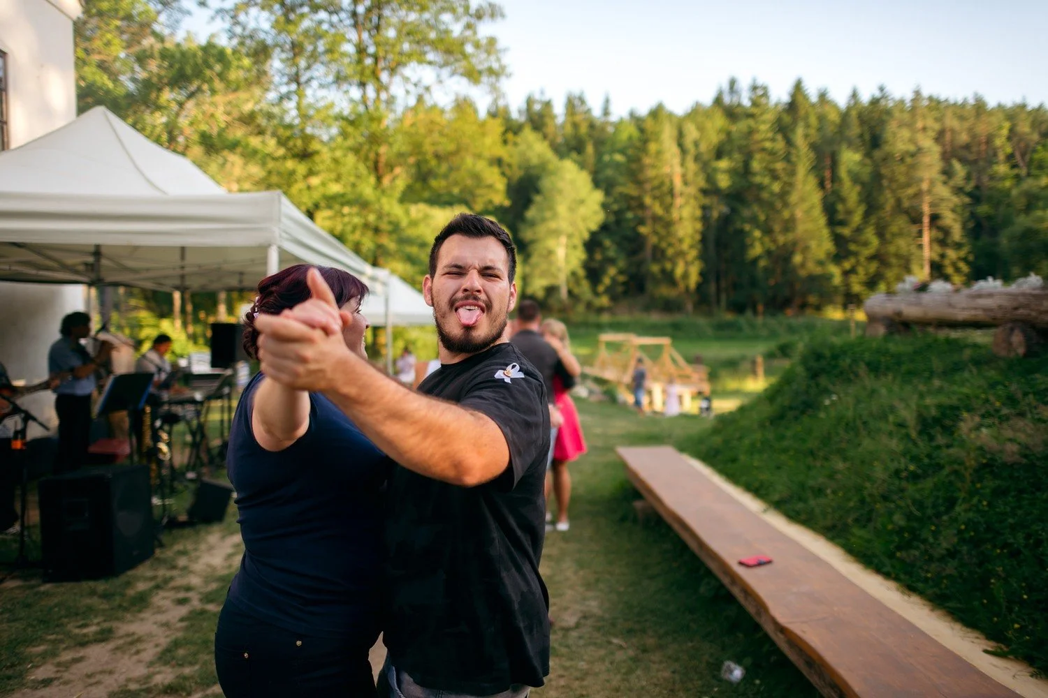 Muž v černém tričku gestikuluje směrem k fotoaparátu u lavičky vedle rybníka.