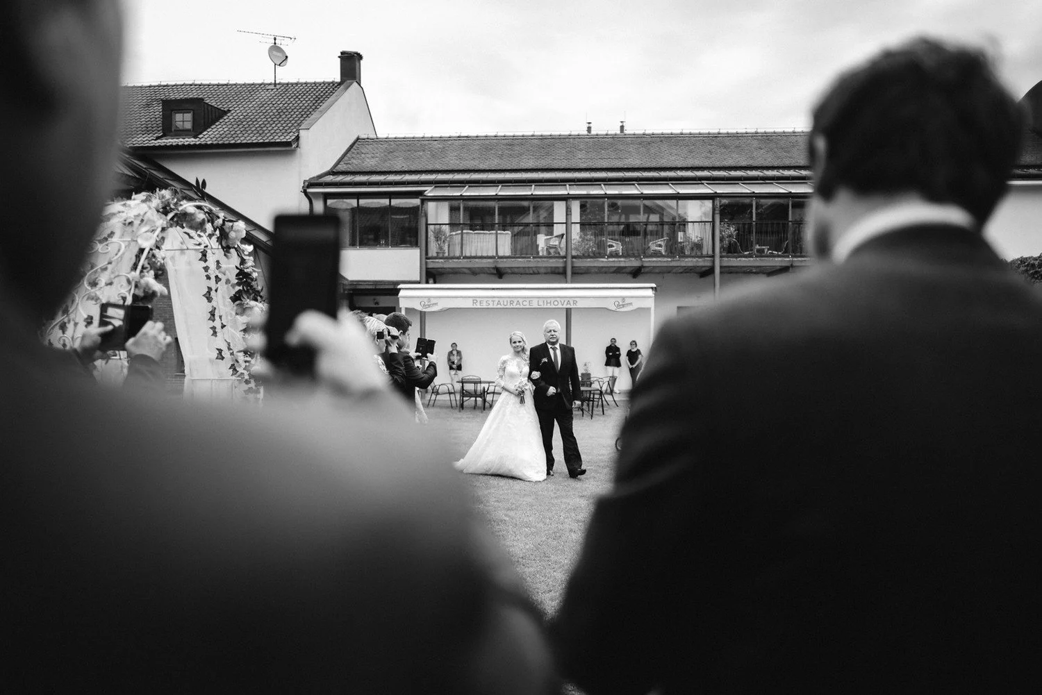 Černobílý pohled mezi rozostřenými postavami zachycuje nevěstu s mužem na protější straně dvora.