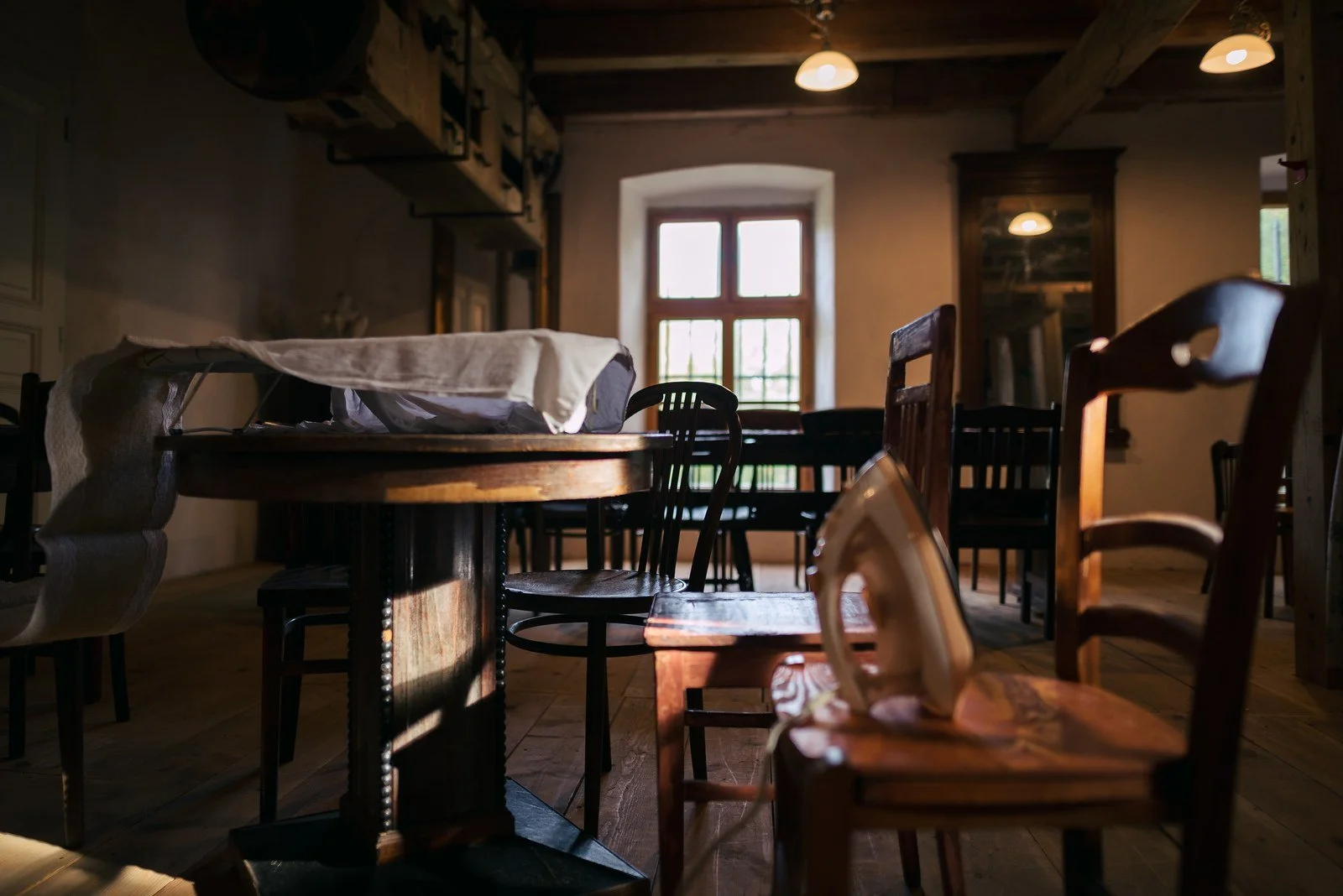 Žehlička leží na dřevěné židli v místnosti se stoly, trámy a oknem.