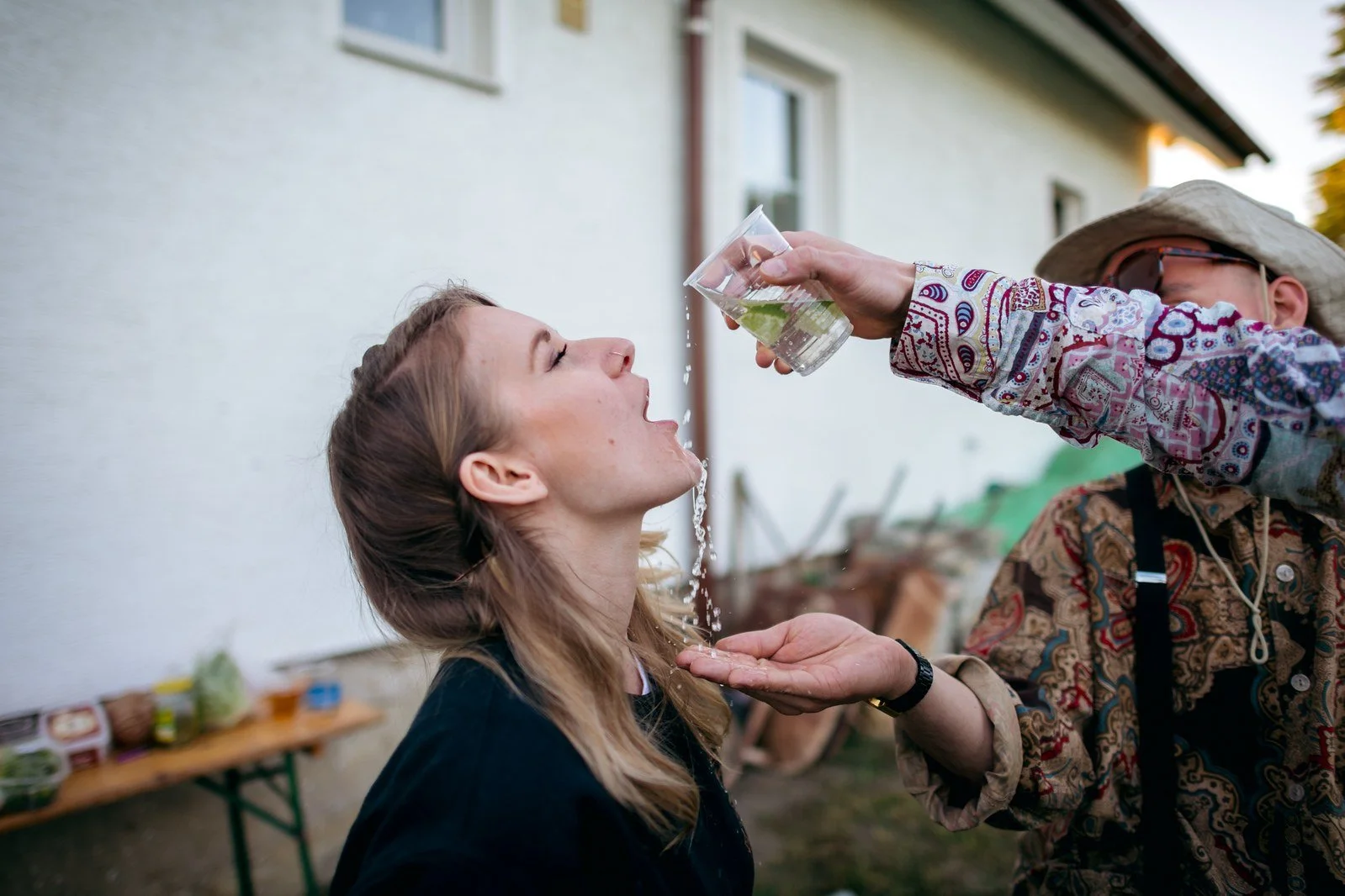Muž v bílé košili pije z lahve při venkovní soutěži mezi hosty.