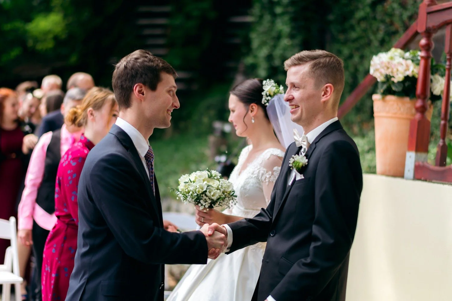 Ženich si podává ruku s mužem v tmavém obleku, nevěsta stojí mezi nimi.