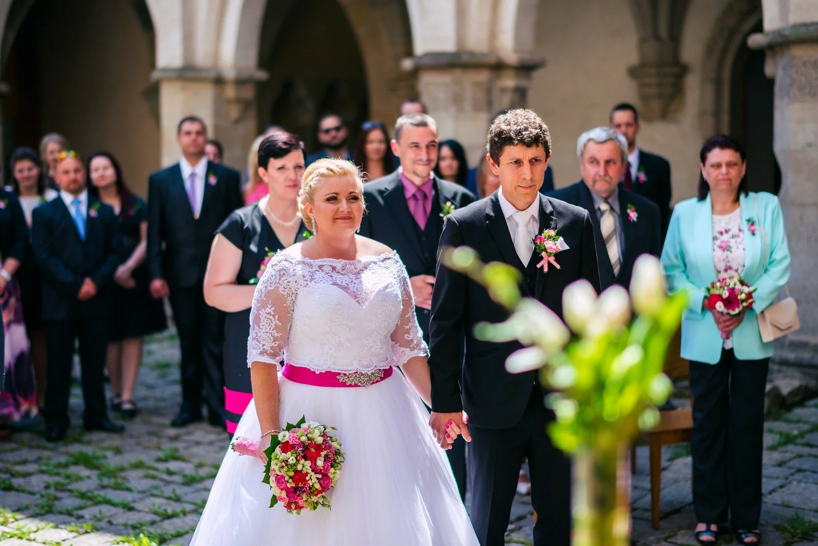 Žena s kyticí a muž stojí čelem mezi soustředěnými hosty.