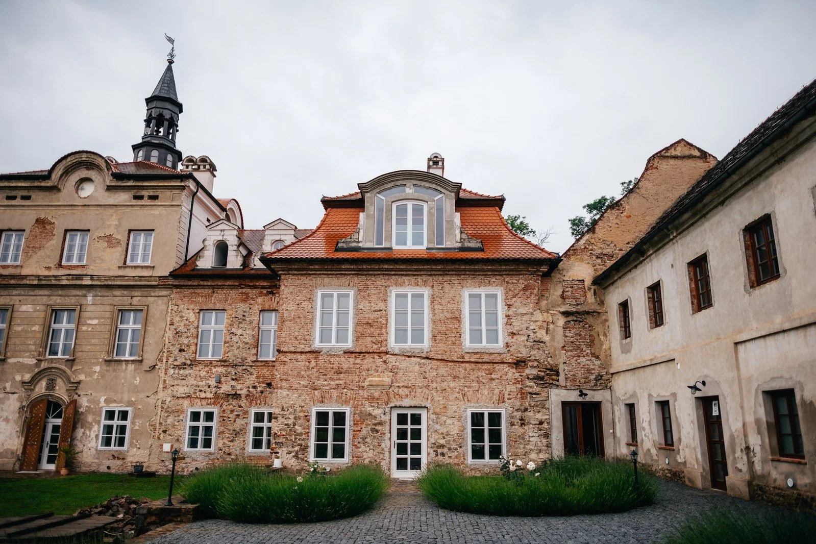 Pohled přes travnatý dvůr zachycuje historickou budovu s kamennými fasádami, řadou oken a věžičkou.