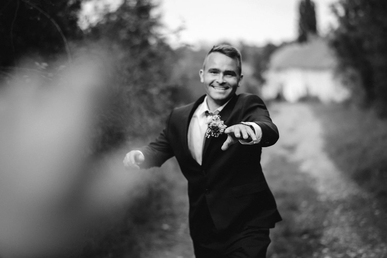 Černobílý portrét usměvavého muže v obleku natahujícího ruku k fotoaparátu.