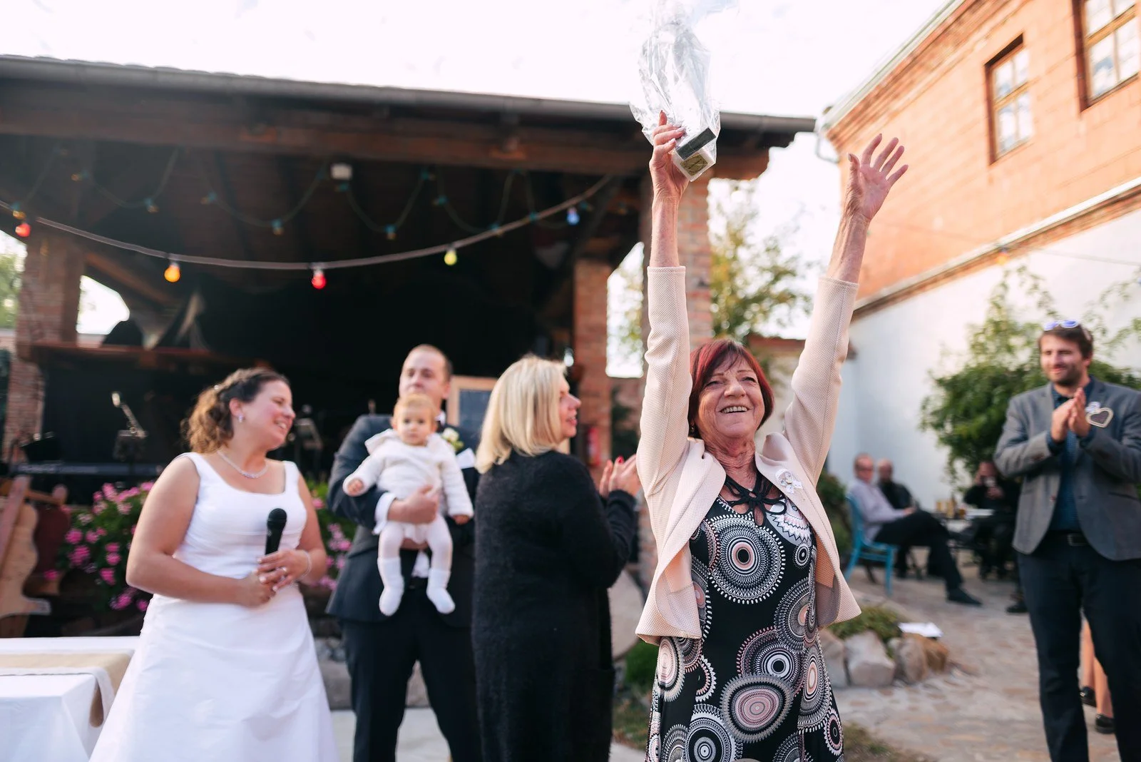 Skupina dospělých slaví na nádvoří; jedna žena drží dítě a jiná zvedá ruku.