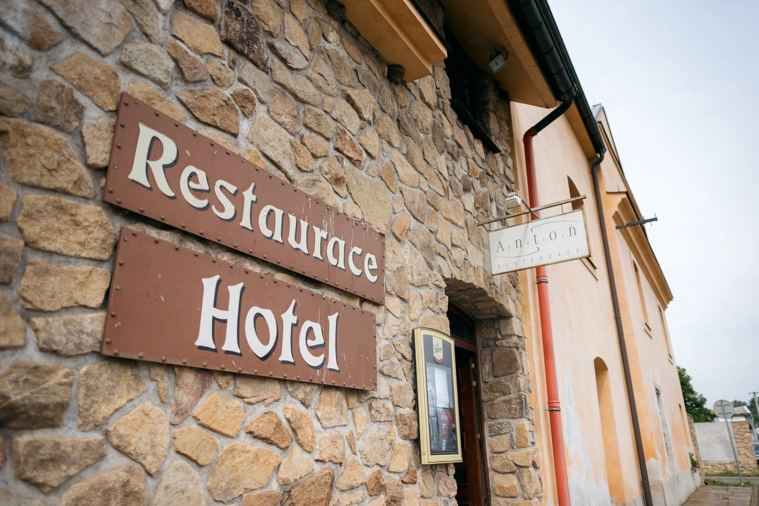 Kamennou fasádu budovy doplňují hnědé cedule s nápisy Restaurace a Hotel.