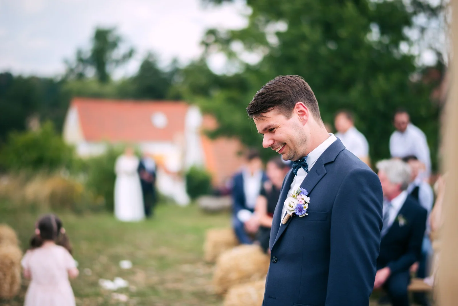 Muž v tmavém obleku se sklopeným pohledem stojí na louce před rozostřenou skupinou lidí.