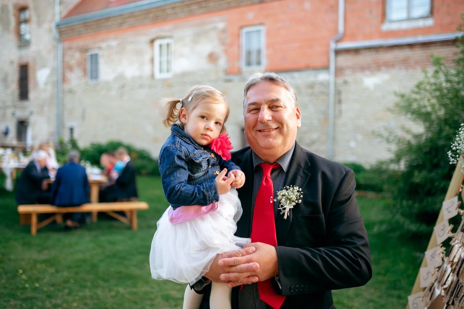 Starší muž drží v náručí malé dítě před cihlovou budovou a zelení.