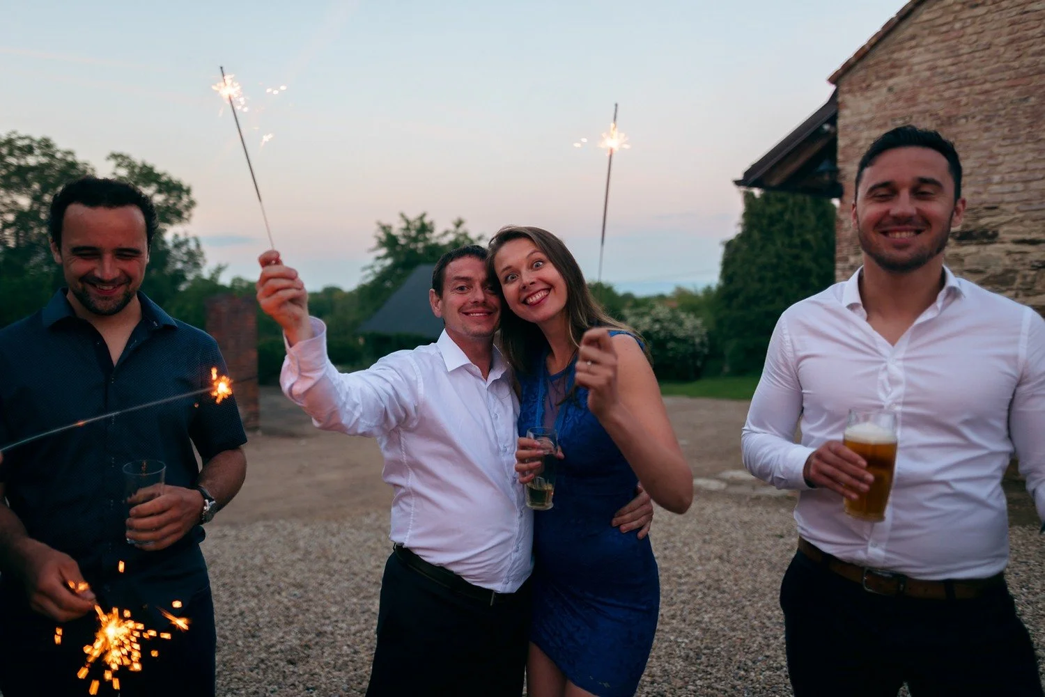 Tři muži a žena pózují venku s hořícími prskavkami a sklenicemi piva.