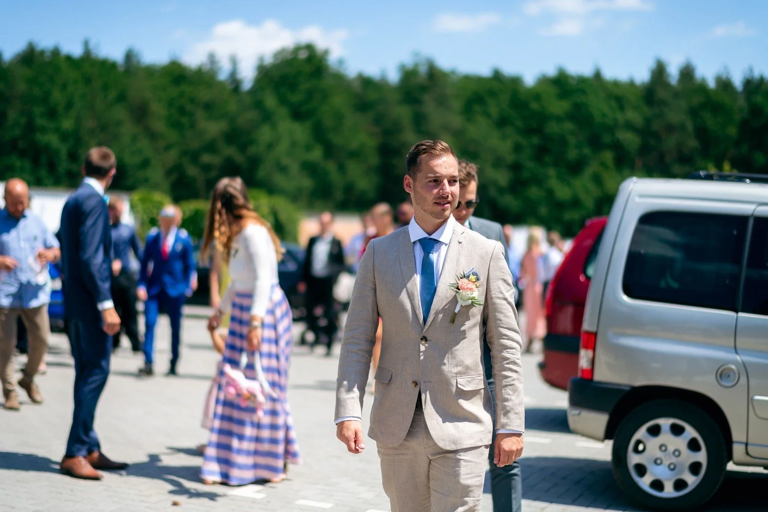 Muž ve světlém obleku s modrou kravatou kráčí venku mezi svatebními hosty.