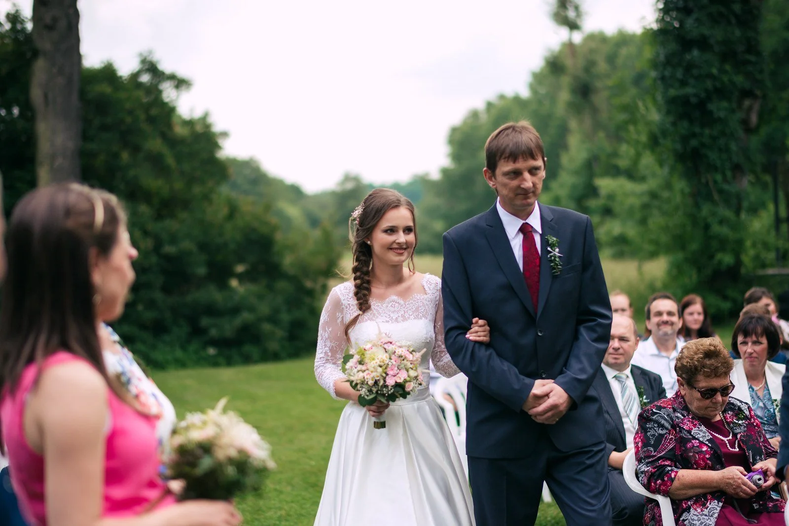Žena s kyticí a muž v obleku přicházejí uličkou mezi hosty.
