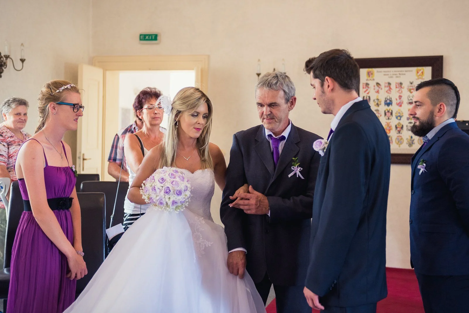 Ivana přichází k obřadu s mužem v tmavém obleku, zatímco Petr čeká vpředu.
