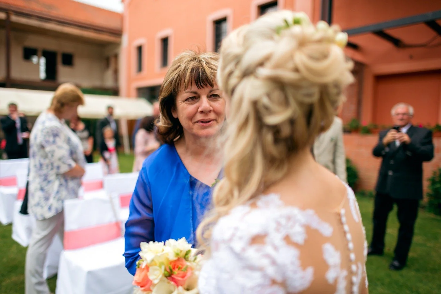 Žena v modrých šatech hovoří s nevěstou, jejíž krajková záda a květinová kytice vyplňují popředí.