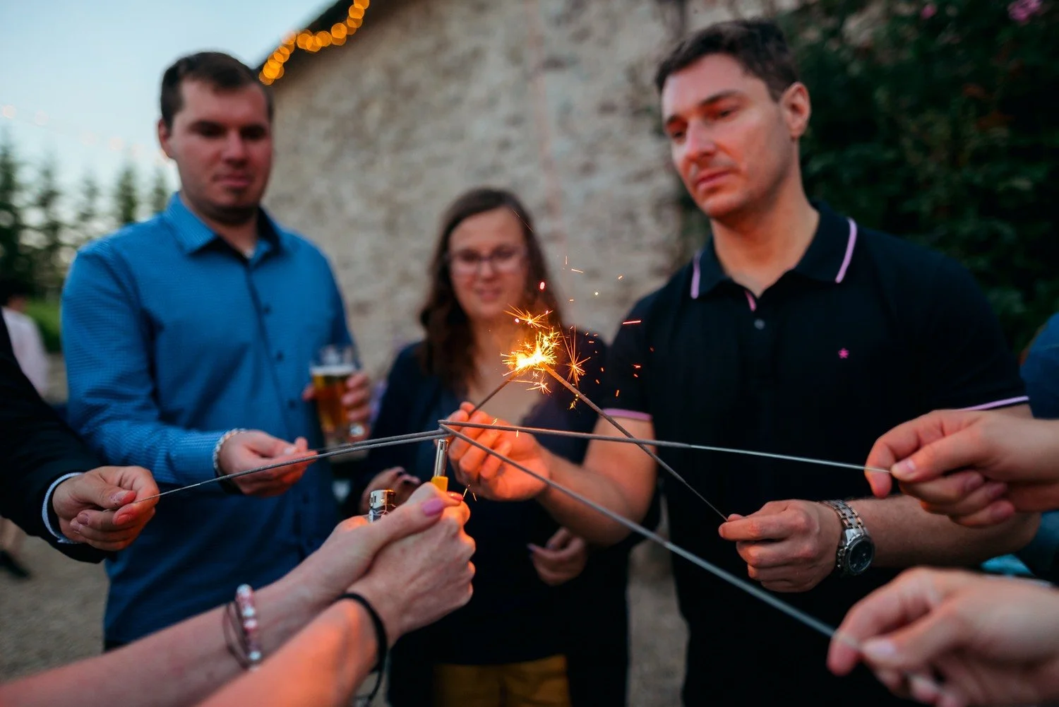 Několik hostů přikládá prskavky k plameni zapalovače na štěrkovém dvoře.