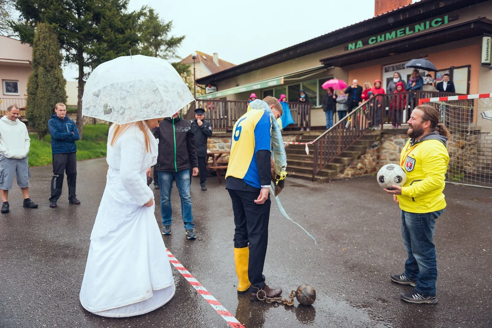 Nevěsta a hosté s deštníky procházejí po deštivé ulici mezi stánky.