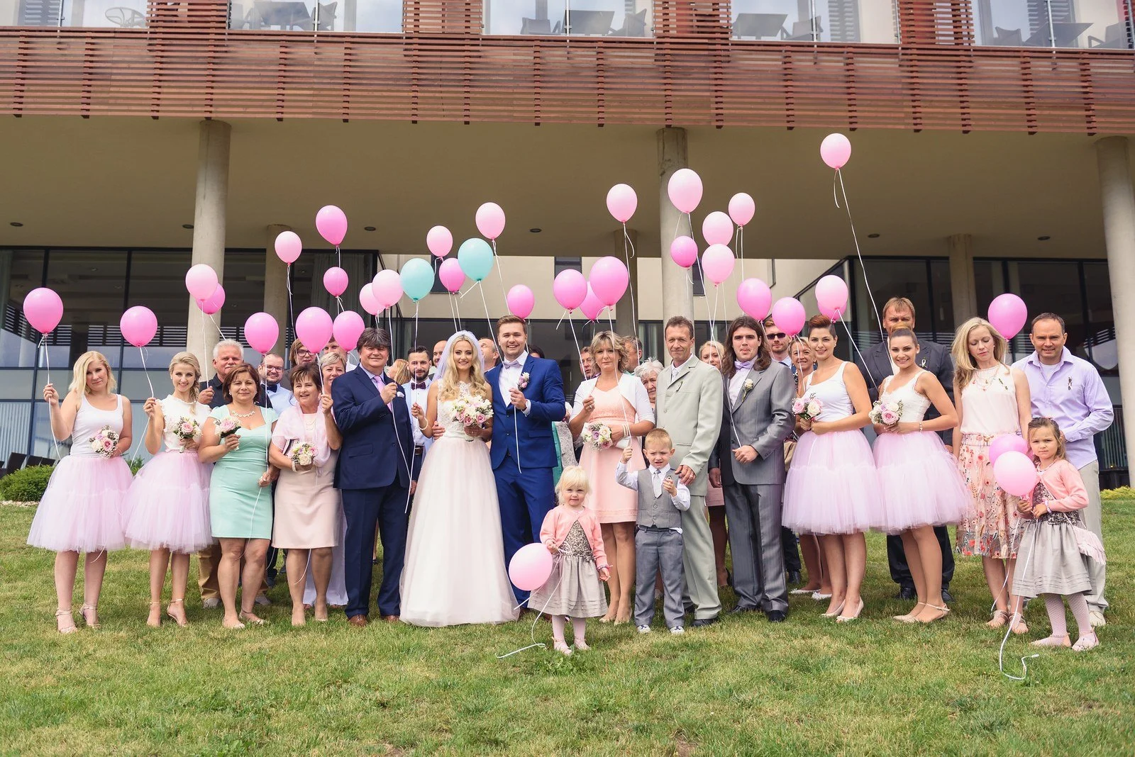 Velká skupina hostů pózuje před budovou s růžovými balonky.