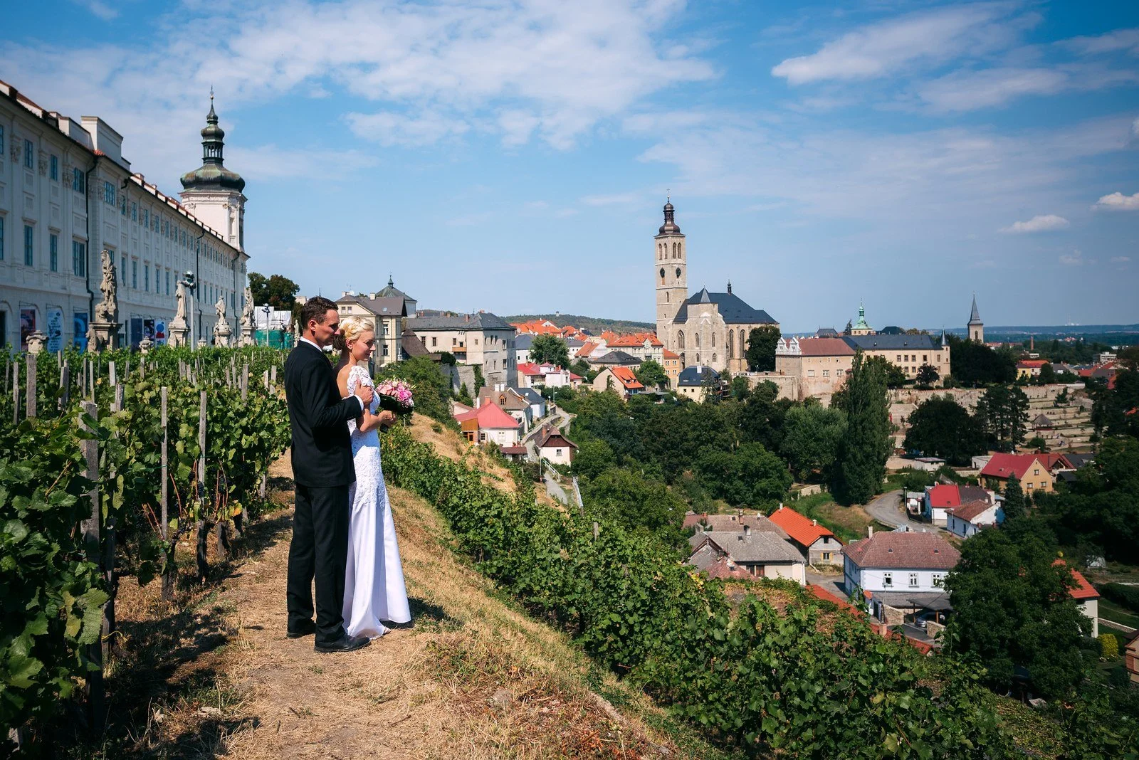 Dvojice stojí na svahu vinice s panoramatem Kutné Hory za sebou.