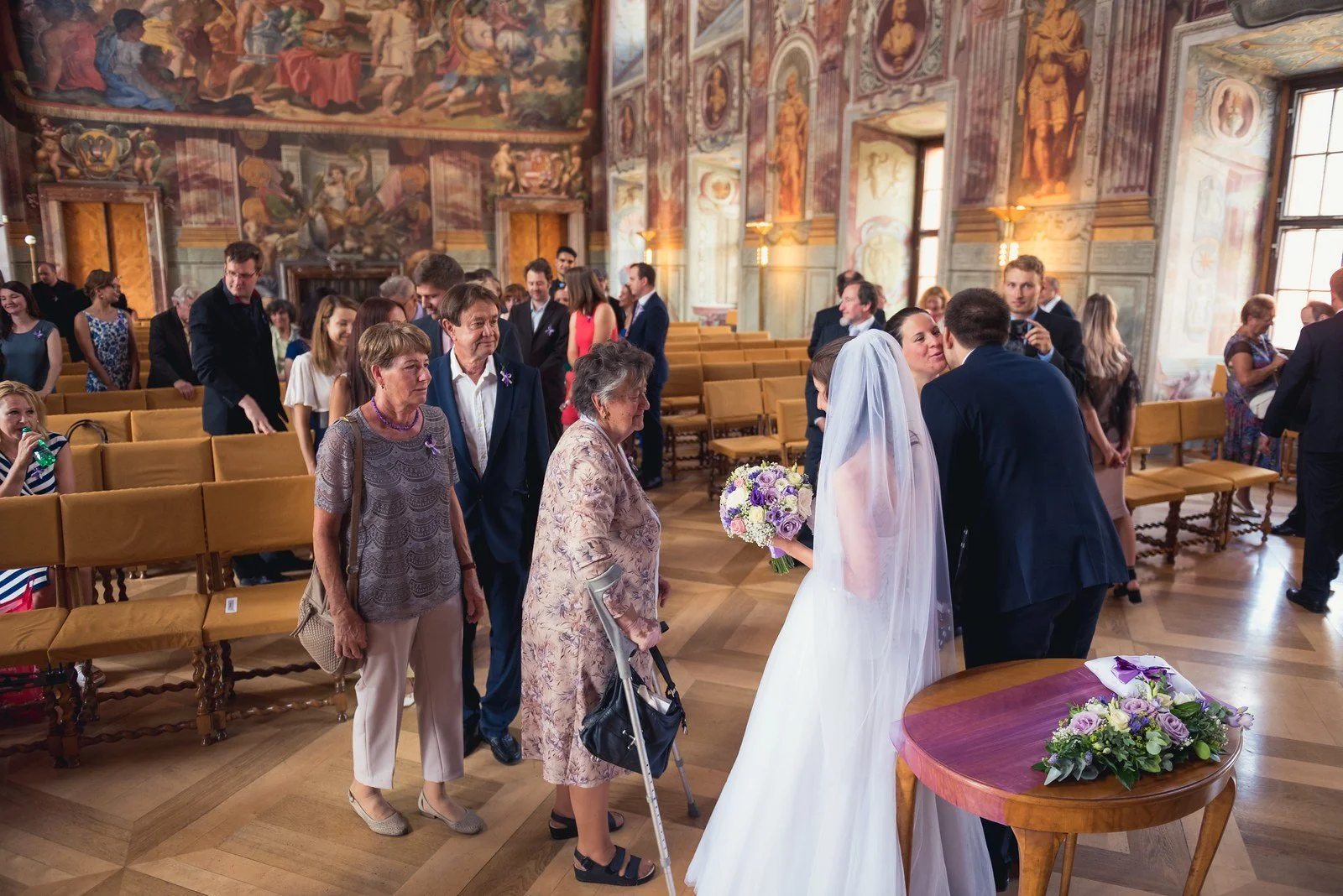 Široký záběr řady lidí při osobních pozdravech zachovává prostorovou orientaci a průběh gratulací.