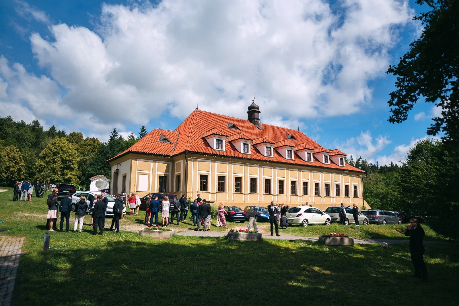 Dvě ženy se soustředí na přípravy u okna, jedna v květovaném oděvu se sklání k druhé.