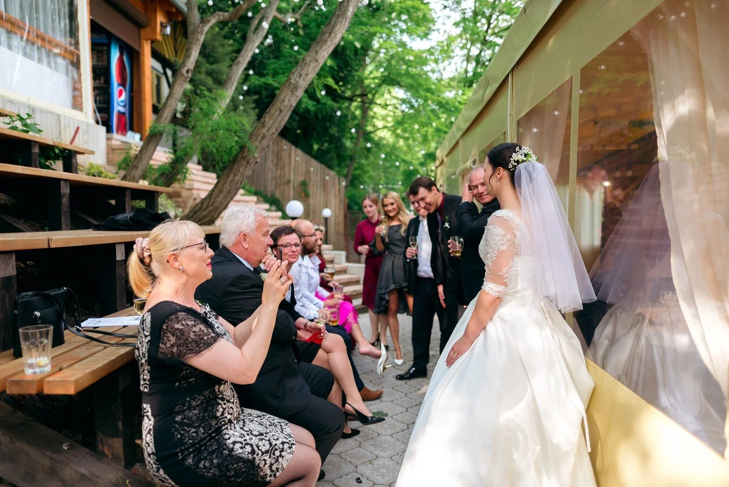 Nevěsta prochází kolem hostů usazených na venkovních dřevěných lavicích.