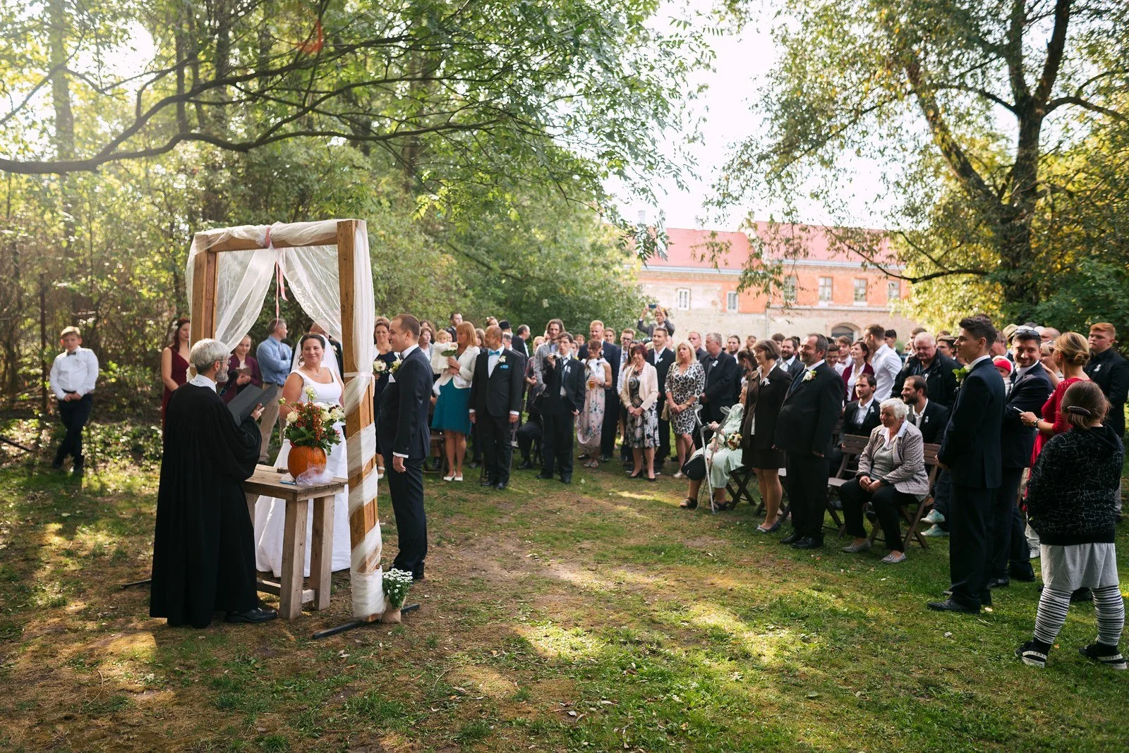 Široký pohled na obřad pod dřevěným rámem obklopený stojícími hosty.
