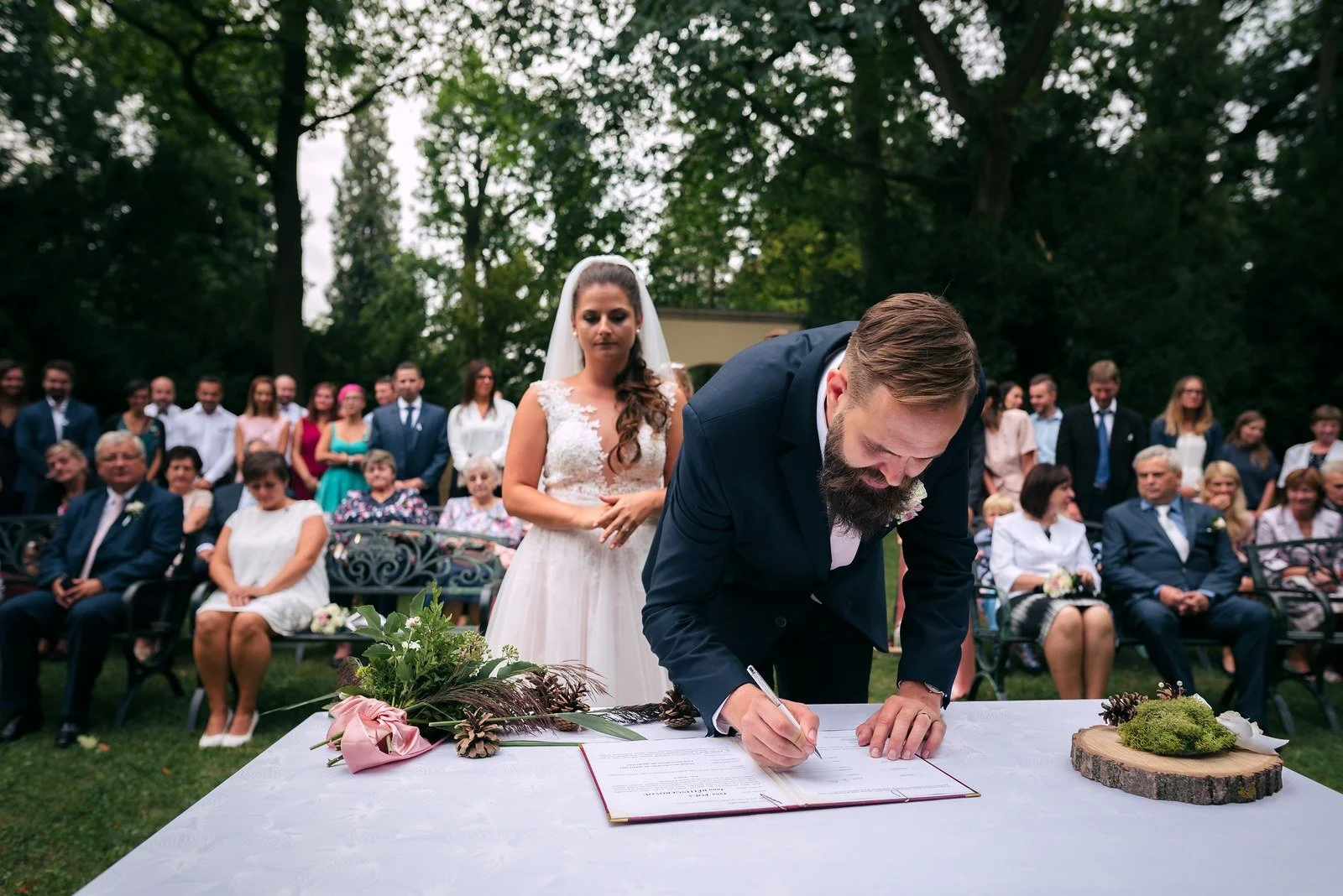Muž v obleku se sklání k podpisu dokumentu na venkovním stolku.