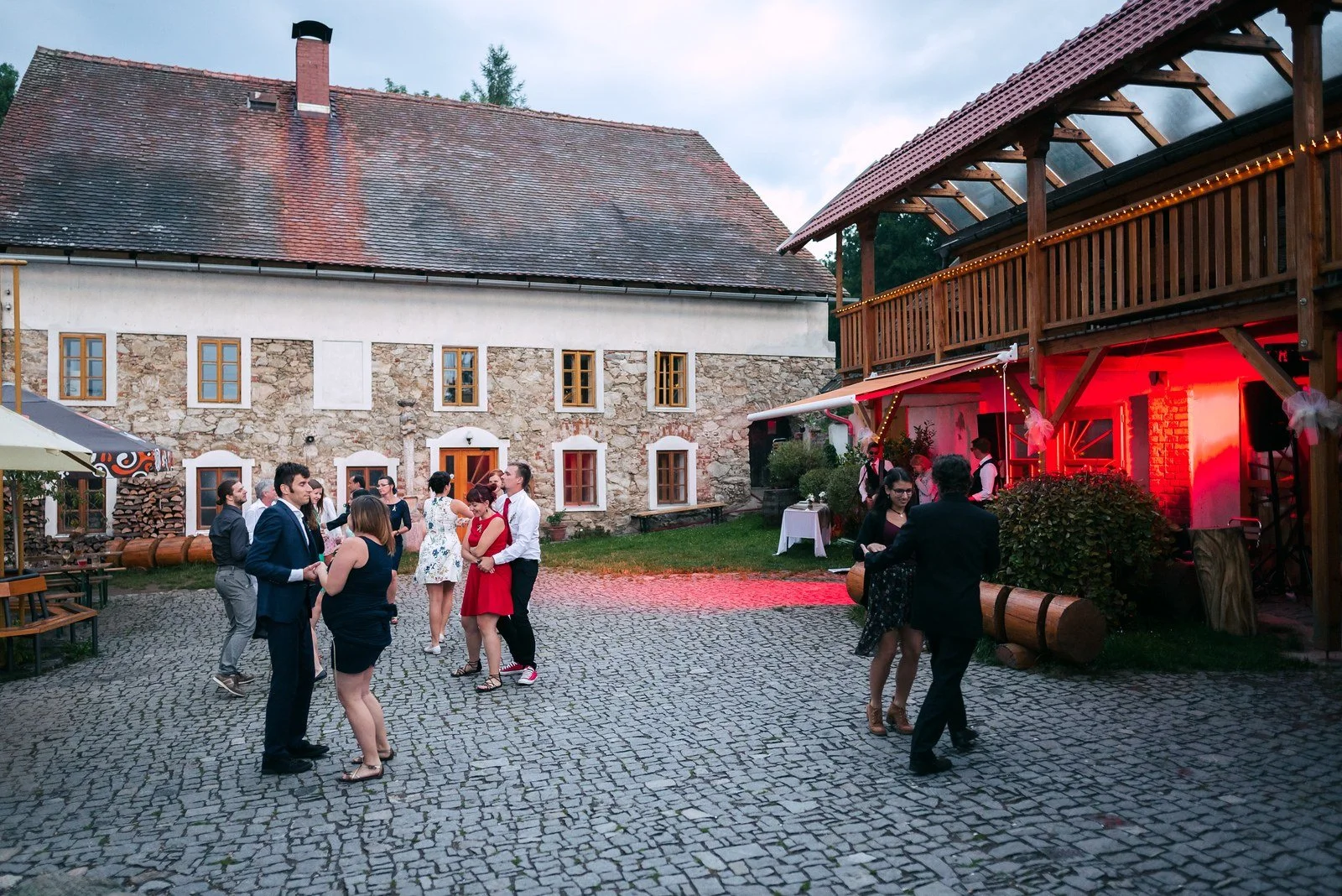 Několik dvojic tančí za soumraku na nádvoří částečně zalitém červeným světlem.