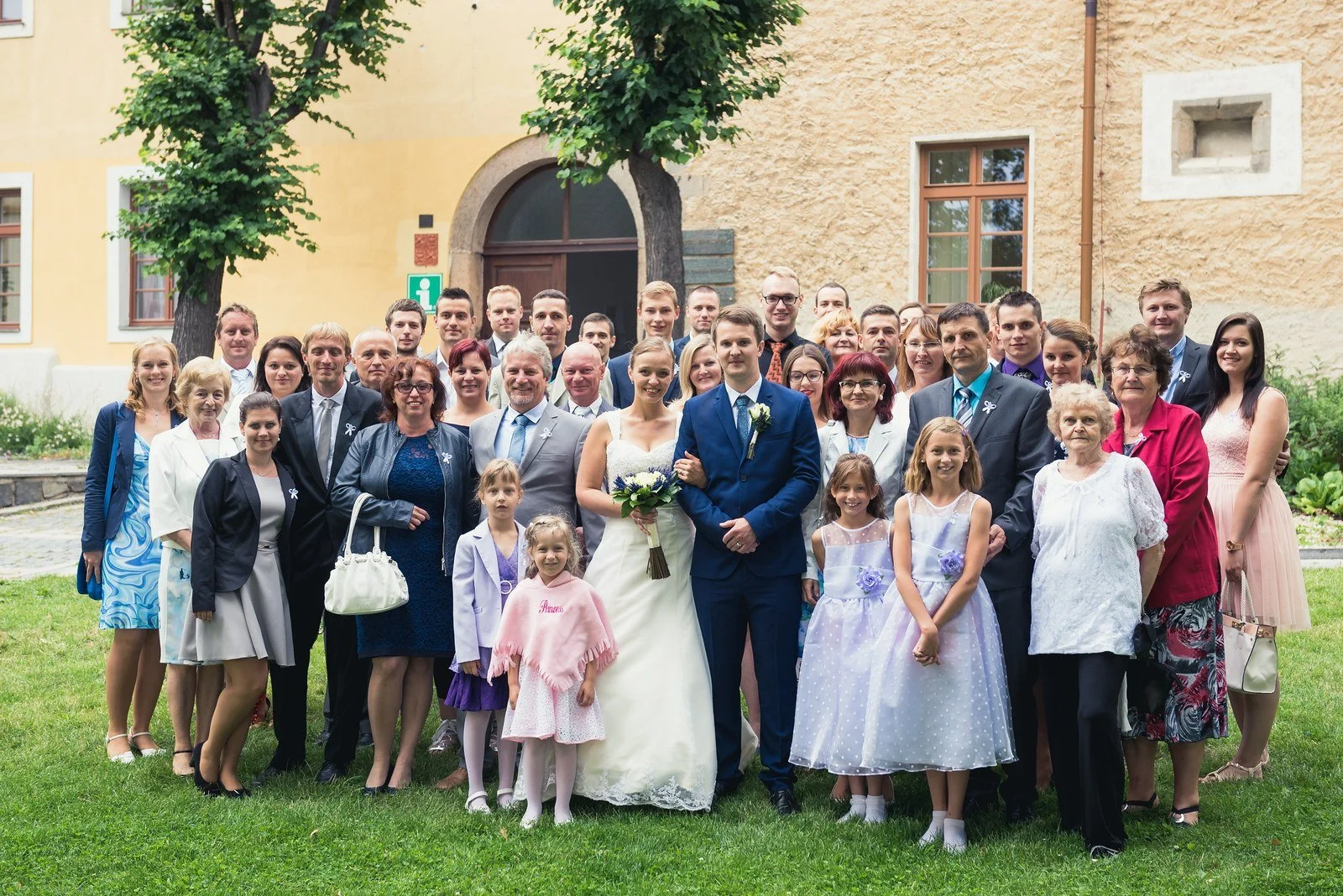 Společná fotografie novomanželů a svatebních hostů před budovou.