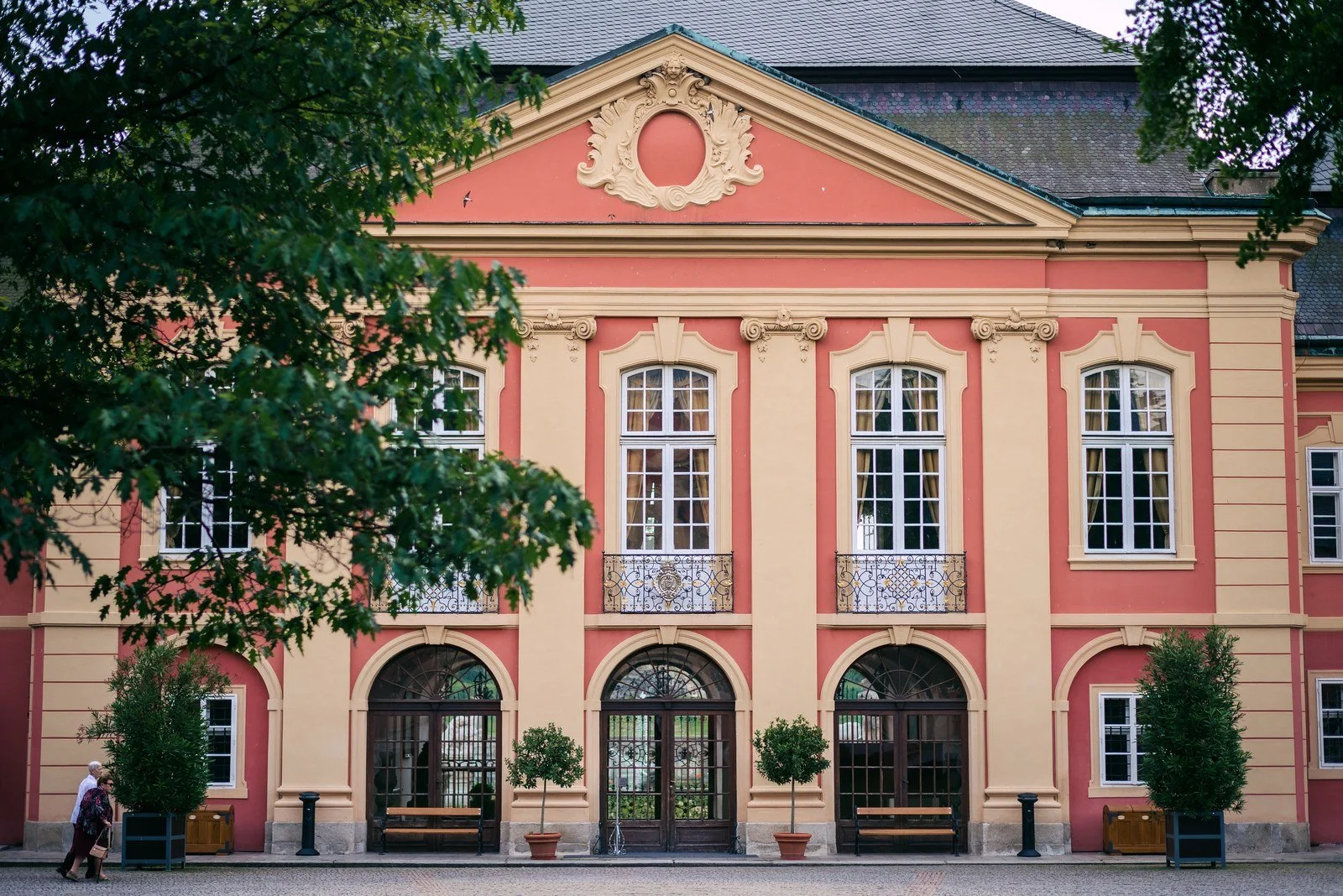 Světle růžová historická budova stojí za upraveným trávníkem.