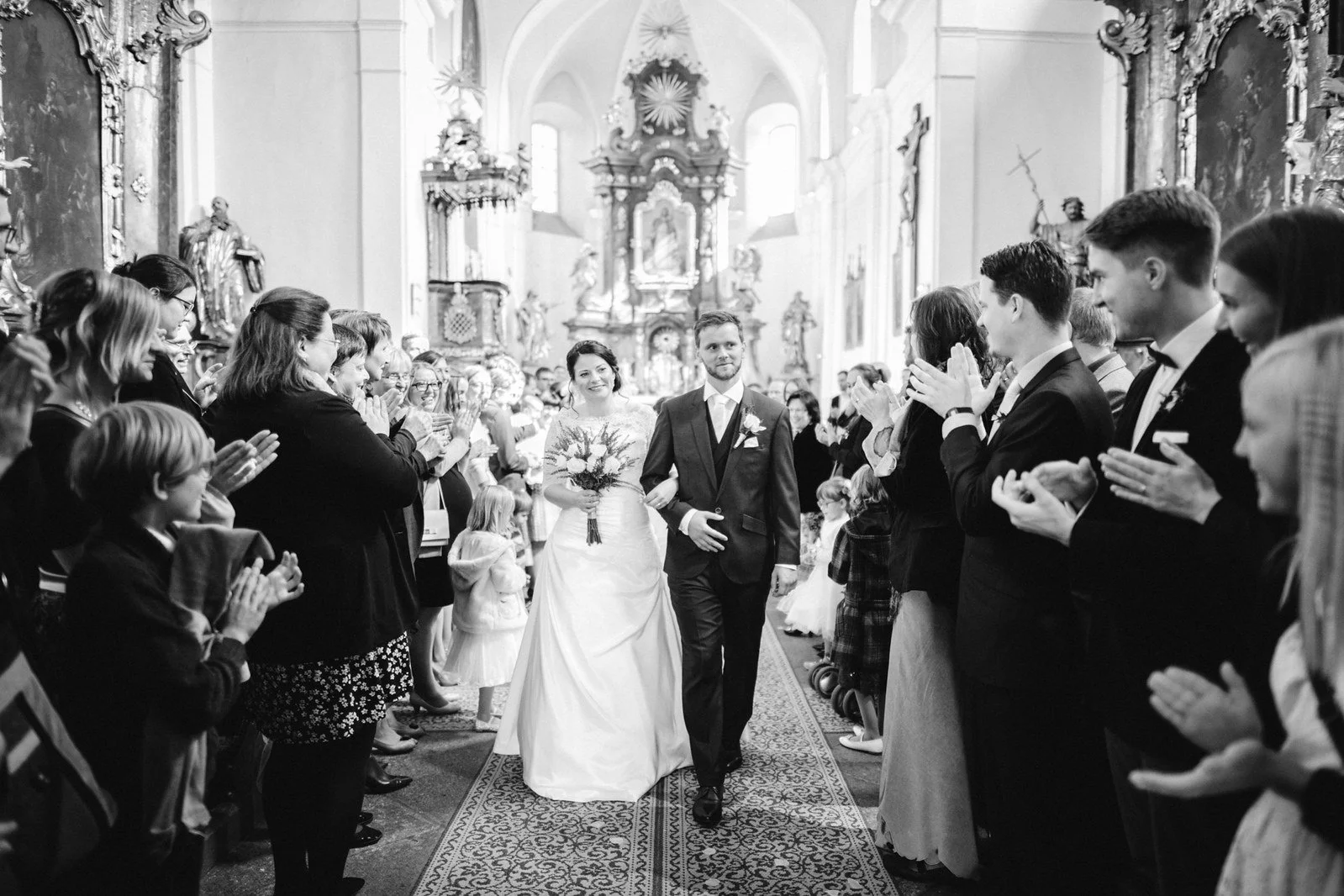 Černobílý široký záběr zachycuje průchod dvojice uličkou mezi řadami tleskajících hostů.