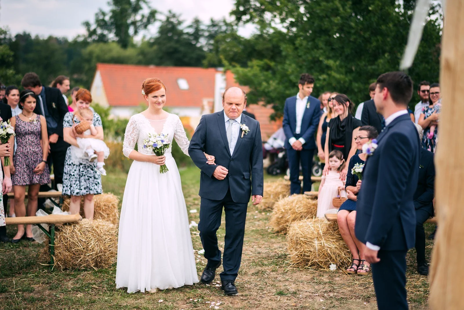 Žena v bílých šatech kráčí po travnaté uličce vedle muže v tmavém obleku.