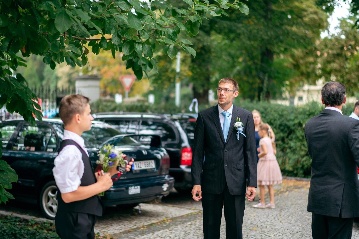 Muži ve společenském oblečení hovoří pod stromy u zaparkovaných aut.